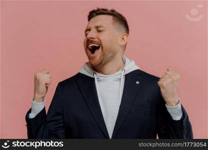 Happy excited young unshaven man gesturing for ecstatic achievement, triumphing and celebrating success, overjoyed redhead guy raising clenched fists and screaming from joy. Winning concept. Happy excited man with closed eyes raising clenched fists and celebrating victory