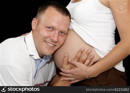 Happy expectant father listening to hear the baby inside his pregnant wife&rsquo;s belly. Black background.