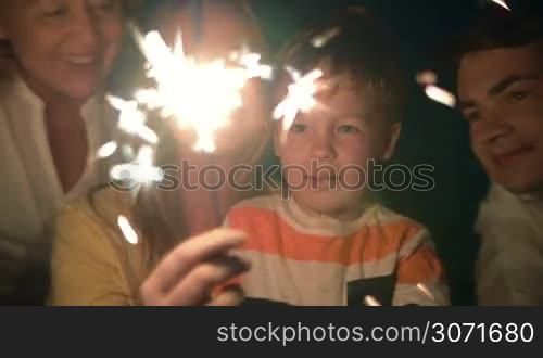Happy family and Bengal fire at night. Mother holding sparkler, then she and father kissing child while he looking at light which going out at the end