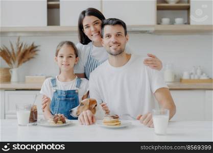 Happy family and dog pose in cozy kitchen, eat fresh homemade pancakes with chocolate and milk, look positively at camera. Mother in apron embraces husband and daughter, likes cooking for them