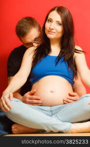 Happy family and waiting for baby concept. Funny man husband hugging his pregnant attractive woman wife,portrait on red