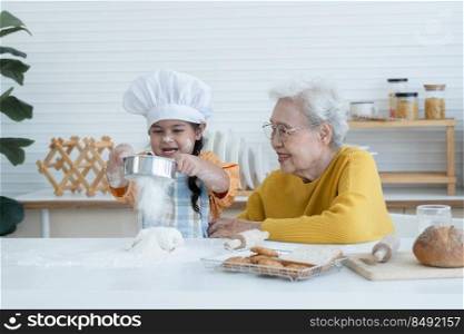Happy family Asian elderly grandmother and little cute grandchild spend time together at kitchen, sift flour on dough for bake cookies or bread, smile and have fun, flour mess up on nose and face