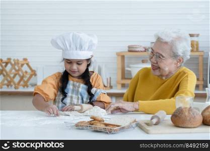 Happy family Asian elderly grandmother and little cute grandchild spend time together at kitchen, knead dough by rolling pin and bake cookies or bread, smile and have fun, flour mess up on face