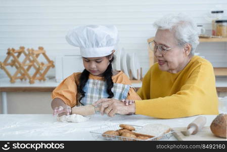 Happy family Asian grandmother and little grandchild spend time together at kitchen, knead dough by rolling pin and bake cookies, elderly grandma teaching and sharing homemade baking skills with niece