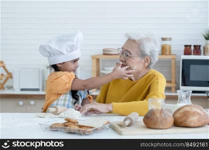 Happy family Asian grandmother and little grandchild spend time together at kitchen, knead dough and bake cookies or bread, niece have fun to tease and put flour on her grandma face at home