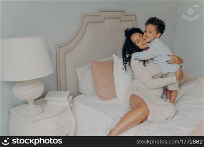 Happy family at home. Smiling pregnant mixed race woman mother in dress with curly hair hugging her beloved sweet little son while sitting on big soft bed. Mom and little child spending time together. Cute little son hugging his pregnant mom, waiting for future brother or sister