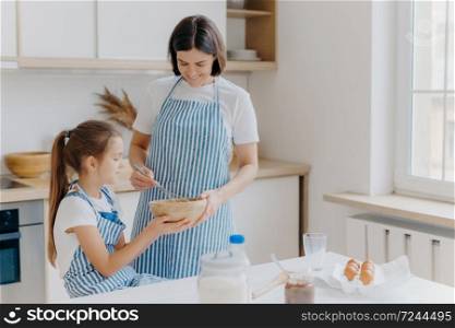Happy family at kitchen. Lovely woman and her daughter prepare bakery together, wear aprons, like cooking together, enjoy domestic atmosphere, have fun indoor. Children, motherhood, baking concept
