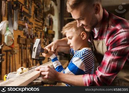 happy family, carpentry, woodwork and people concept - father and little son with hammer hammering nail into wood plank at workshop