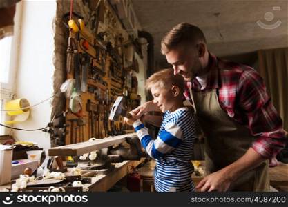 happy family, carpentry, woodwork and people concept - father and little son with hammer hammering nail into wood plank at workshop