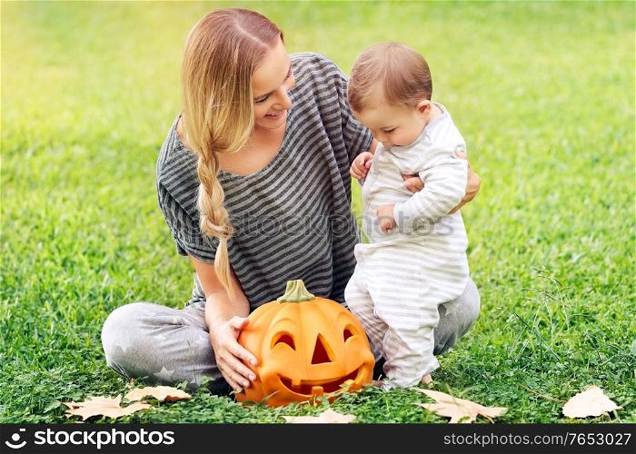 Happy family celebrating Halloween outdoors, mother with cute little baby boy playing with carved pumpkin on the green field, enjoying autumn holiday