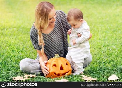 Happy family celebrating Halloween outdoors, mother with cute little baby boy playing with carved pumpkin on the green field, enjoying autumn holiday