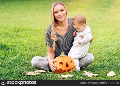 Happy family celebrating Halloween outdoors, mother with cute little baby boy playing with carved pumpkin on the grass field, enjoying autumn holiday