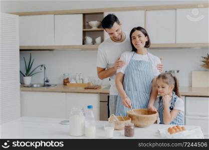Happy family cook together at kitchen. Father, mother and dauther busy preparing delicious meal at home. Husband embraces wife who whisks and prepares dough, bake cookies. Food, togetherness