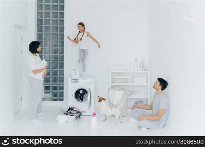 Happy family do laundry at home, father sits on floor in lotus pose, mother stands with white towel, look at child who dances happily on washing machine, pedigree dog near. Household chores.