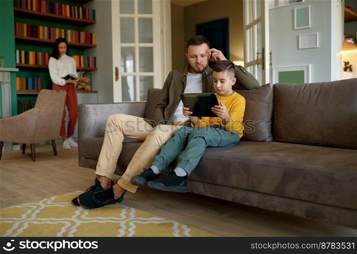 Happy family enjoy fun time together on weekend at home. Father and son embracing while using mobile tablet while mother reading book. Happy father and son embracing while using mobile tablet at home