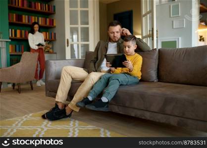 Happy family enjoy fun time together on weekend at home. Father and son embracing while using mobile tablet while mother reading book. Happy father and son embracing while using mobile tablet at home