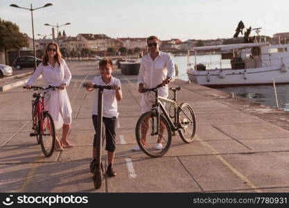 Happy family enjoying a beautiful morning by the sea together, parents riding a bike and their son riding an electric scooter. Selective focus. High-quality photo. Happy family enjoying a beautiful morning by the sea together, parents riding a bike and their son riding an electric scooter. Selective focus 