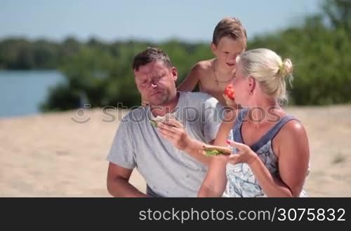 Happy family enjoying lunch on the beach during picnic on summer vacation. Son eating tomato and kissing his parents