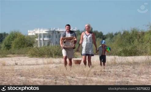 Happy family going on picnic to the beach. Father carrying a picnic basket. Mother with basket of fresh fruits holding son&acute;s hand.