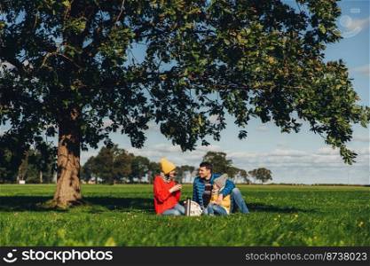 Happy family have autumn picnic, sit on green grass, drink hot tea, communicate with each other, enjoy calm atmosphere, admire blue sky and beautiful landscapes. Friendly family members together