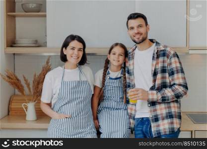 Happy family have great time to≥ther, pose in modern kitchen at home. Glad man in checkered shirt holds glass of juice, smallχld withπ>ails, pretty housewife in apron. Girl with parents