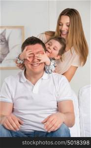 Happy family having fun in the living room: son surprisng his father by covering his eyes
