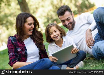 Happy family in a urban park playing with tablet computer. Father, mother and little daughter sitting on the grass laughing.