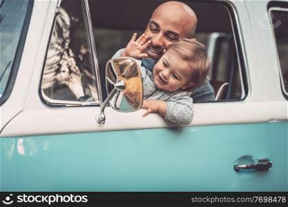 Happy family in the car, summer travel in the van, cheerful father with his adorable little son driving wagon, smiling and waving his hands, enjoying parenthood