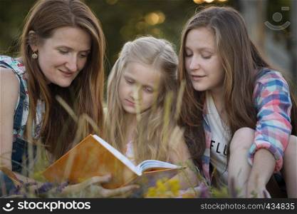 Happy family - mom and two daughters are sitting in a meadow and reading a book
