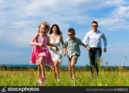 Happy family - mother, father, children - running over a green meadow in summer kicking a soccer ball