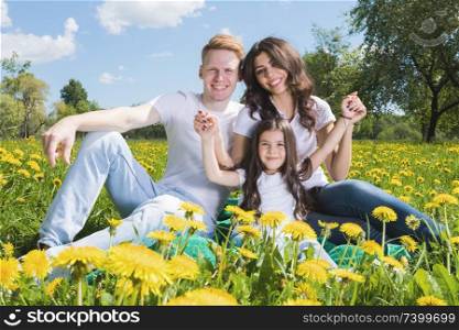 Happy family of parents and daughter sitting on a meadow of dandelion flowers and smiling. Family on flowers meadow