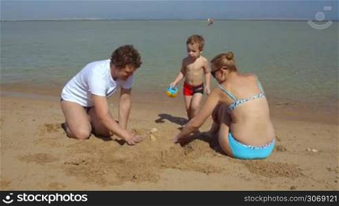 Happy family of three on the beach. Mother and father making sand castle, their son bringing water
