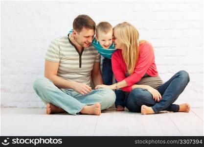 Happy family of three sitting on the floor near the wall and laughing: mother, father and little boy. Mother is pregnant.