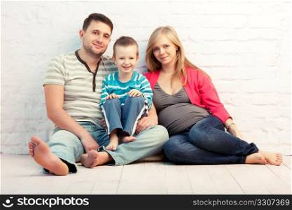 Happy family of three sitting on the floor near the wall: mother, father and little boy. Mother is pregnant.