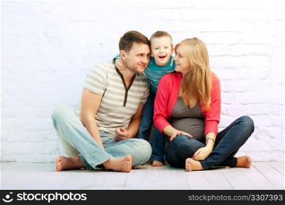 Happy family of three sitting on the floor near the wall: mother, father and little boy. Mother is pregnant. Son is wondered.