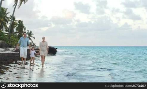 Happy family of three walking along the shore in tropics. They holding hands, water washing their feet