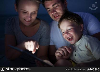 Happy family of three with pad late in the evening. Laughing child looking at screen where mother pointing at something