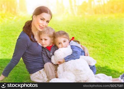 Happy family outdoors, young mother with two precious child sitting of fresh green grass field, summer vacation, love and togetherness concept