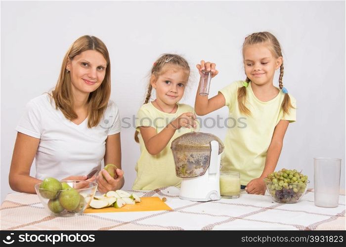Happy family prepares freshly squeezed juice in a juicer. Young beautiful mother and two daughters sitting at a table squeezed juice from pears and grapes with a juicer