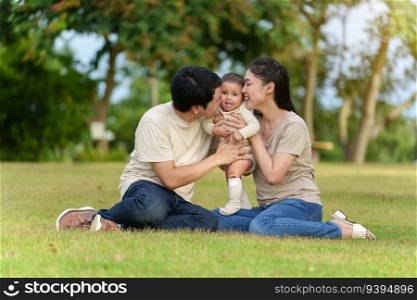 happy family sitting on a green grass filed. father and mother kissing their infand baby in the park