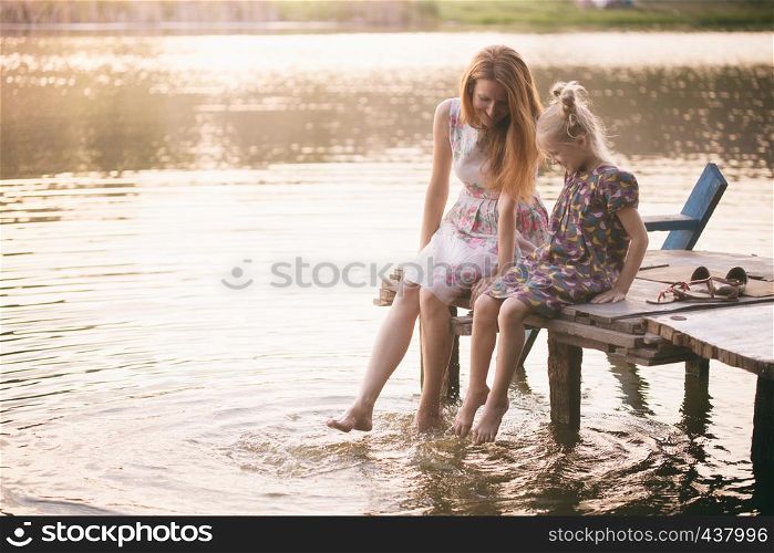 Happy family - smiling mother and daughter sitting on the pier warm autumn day dangling his legs in the water and splash in the foreground