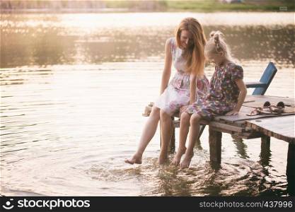Happy family - smiling mother and daughter sitting on the pier warm autumn day dangling his legs in the water and splash in the foreground