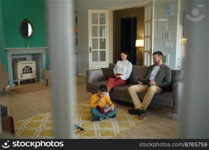 Happy family spending time at home together in evening. Father and mother using digital gadget, little son reading book sitting on floor. Happy family spending time at home together in evening