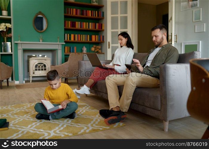 Happy family spending time at home together in evening. Father and mother using digital gadget, little son reading book sitting on floor. Happy family spending time at home together in evening