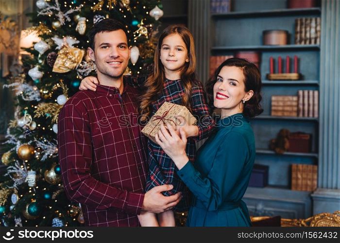 Happy family stand together against decorated fir tree, look happily into camera, hold present, celebrate New Year in family circle. Small girl being glad to spend holidays with her mother and father