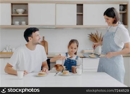 Happy family time and breakfast concept. Cheerful wife and mother prepares delicious pancakes for family members, father, daughter and dog enjoy eating and tasting dessert at home, add chocolate