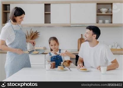 Happy family time and breakfast concept. Cheerful wife and mother prepares delicious pancakes for family members, father, daughter and dog enjoy eating and tasting dessert at home, add chocolate