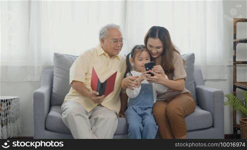 Happy family time relax. Asian grandparents laughing taking selfie with granddaughter on sofa at home by mobile smartphone. senior man or grandpa, daughter and girl kid in living room together