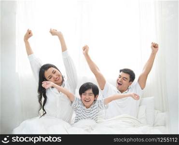 Happy family wake up in morning stretching hand rise up to the air while sitting on bed in bedroom with big window in background.Caucasian young man with asian woman and little boy exercise on bed.