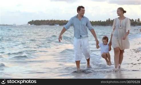 Happy family walking along the shore in tropics. They holding hands, water washing their feet